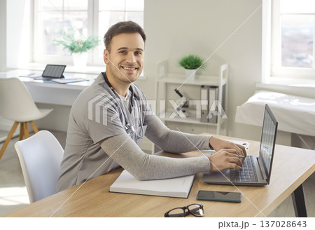 Smiling friendly doctor keyboarding on laptop computer, writing in notebook and consulting internet 137028643