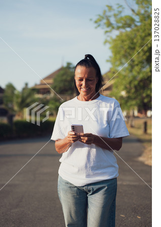 Smiling Mature Scandinavian Tourist Woman Walking Outdoors on Summer Holiday Texting With Phone 137028825