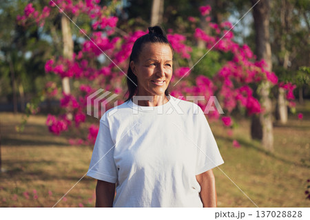 Smiling Mature Woman Standing in Tropical Garden with Pink Flowers Smiling Mature Woman Standing in Tropical Garden with Pink Flowers 137028828