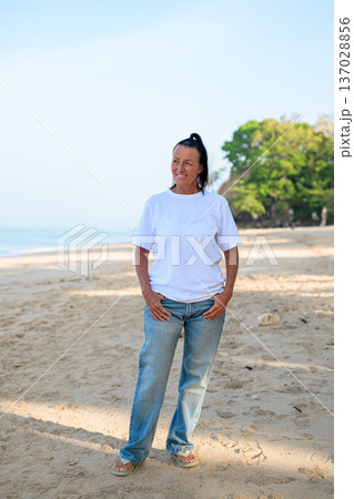 Mature woman standing on tropical beach wearing 90s style normcore fashion jeans and white t-shirt 137028856