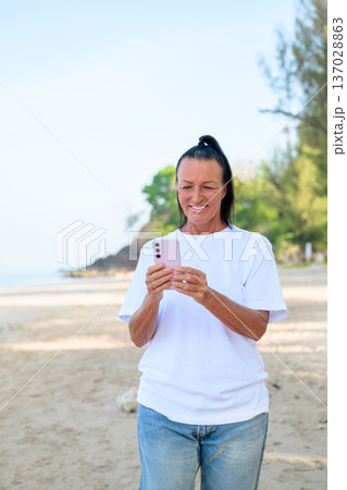 Mature woman standing on tropical beach wearing 90s style normcore fashion jeans and white t-shirt Mature woman standing on tropical beach wearing 90s style normcore fashion jeans and white t-shirt 137028863