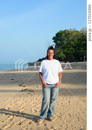 Mature woman standing on tropical beach wearing 90s style normcore fashion jeans and white t-shirt Mature woman standing on tropical beach wearing 90s style normcore fashion jeans and white t-shirt 137028866