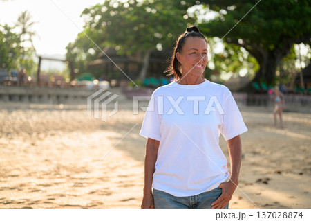 Mature woman standing on tropical beach wearing 90s style normcore fashion jeans and white t-shirt Mature woman standing on tropical beach wearing 90s style normcore fashion jeans and white t-shirt 137028874