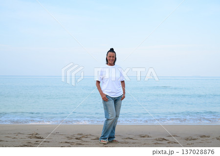 Mature woman standing on tropical beach wearing 90s style normcore fashion jeans and white t-shirt Mature woman standing on tropical beach wearing 90s style normcore fashion jeans and white t-shirt 137028876