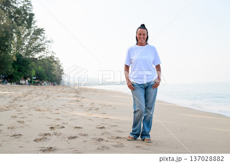 Mature woman standing on tropical beach wearing 90s style normcore fashion jeans and white t-shirt Mature woman standing on tropical beach wearing 90s style normcore fashion jeans and white t-shirt 137028882