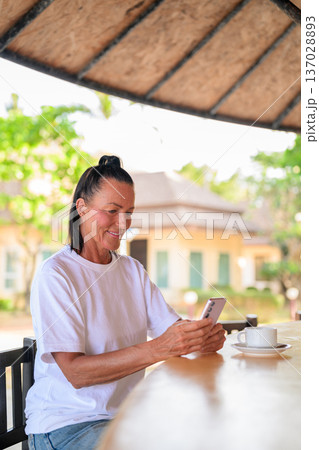 Senior woman texting on phone while enjoying coffee in tropical setting Senior woman texting on phone while enjoying coffee in tropical setting 137028893