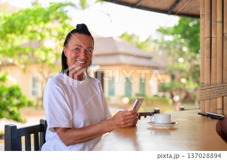 Senior woman texting on phone while enjoying coffee in tropical setting 137028894