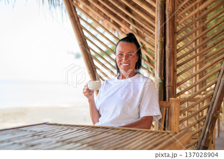 Smiling Mature Woman Drinking Coffee on Beachside Bamboo Terrace Smiling Mature Woman Drinking Coffee on Beachside Bamboo Terrace 137028904
