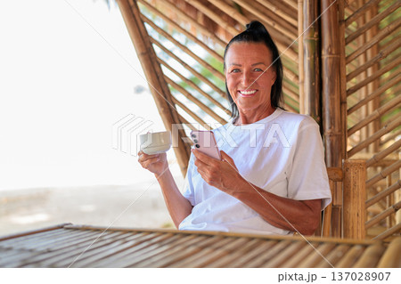Smiling Senior Woman Using Smartphone and Drinking Coffee on Tropical Terrace Smiling Senior Woman Using Smartphone and Drinking Coffee on Tropical Terrace 137028907