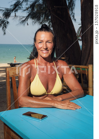 Confident senior woman in bikini relaxing at seaside cafe table with smartphone 137028918