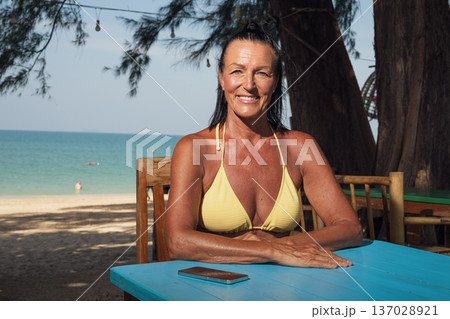 Confident senior woman in bikini relaxing at seaside cafe table with smartphone 137028921