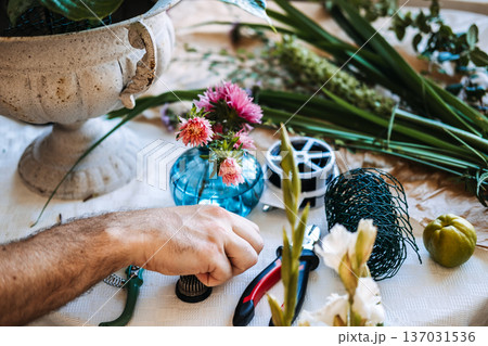 Man places small flower frog on table next to floral tools and aster blooms. Artisanal floral design, master florist at work, handcrafted aesthetic, boutique floral studio. 137031536