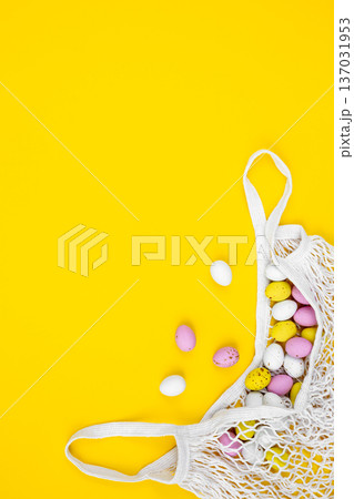 Colored Easter eggs in a mesh bag on a yellow background, flat lay. 137031953