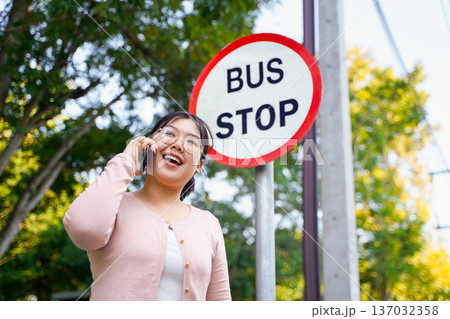 Young Asian woman talking on a smartphone while waiting at a bus stop. Young Asian woman talking on a smartphone while waiting at a bus stop. 137032358