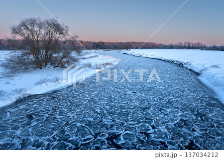 Beautiful nature landscape of winter river with cracked ice at sunset. Warm golden light reflects off the frozen surface, with a dark forest silhouette on the horizon. 137034212