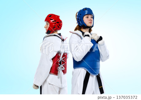 Female taekwondo athletes standing back to back in red and blue protective armor on light blue background. 137035372