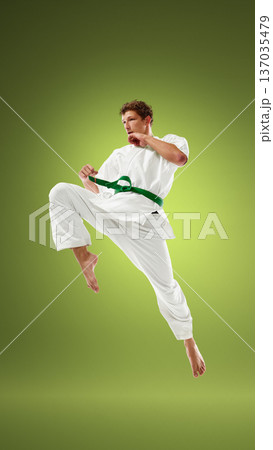 Young martial artist executing high knee strike in white karate uniform with green belt. 137035479