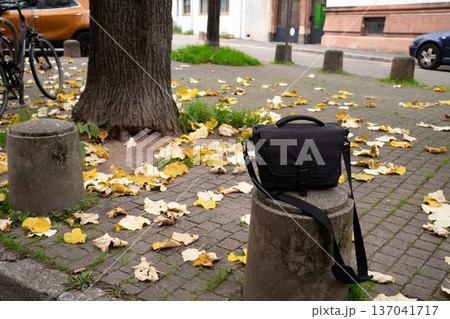 Black camera bag resting on a stone parking road block. Everyday urban scene showing a personal accessory, representing photography, mobility, and city life. Black camera bag resting on a stone parking road block. Everyday urban scene showing a personal accessory, representing photography, mobility, and city life. 137041717