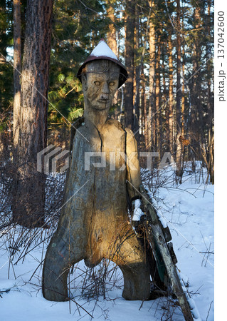 Wooden man statue in snow with a hat in a winter forest. Rustic art and traditional carving concept for landscape design element. Wooden man statue in snow with a hat in a winter forest. Rustic art and traditional carving concept for landscape design element. 137042600