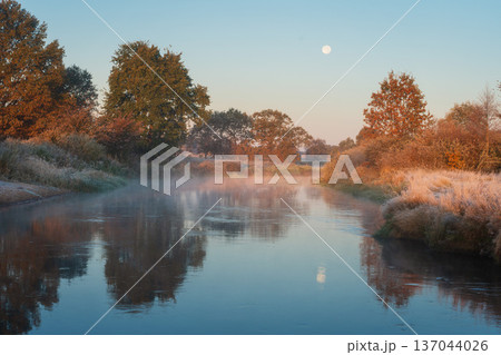A calm autumn morning on a misty river. Wild trees with colorful foliage line the banks under the soft glow of the early sun and a fading colorful moon. A calm autumn morning on a misty river. Wild trees with colorful foliage line the banks under the soft glow of the early sun and a fading colorful moon. 137044026