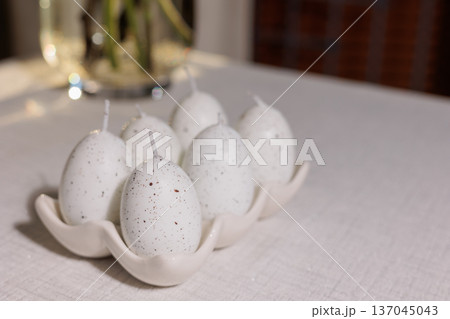 White speckled egg shaped candles standing in a cream ceramic holder on a white table, providing festive decor for an easter celebration or spring gathering White speckled egg shaped candles standing in a cream ceramic holder on a white table, providing festive decor for an easter celebration or spring gathering 137045043