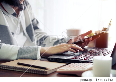 Woman's hand holding a smartphone and using a laptop computer to search for information. at the comfort of home office On a wooden table with a bright white curtain as background 137045148