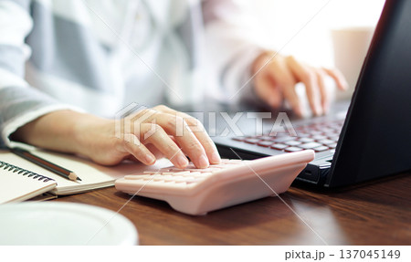 close up woman's hand pressing calculator, calculating data obtained from computer notebook on wooden desk in a bright and comfortable office 137045149