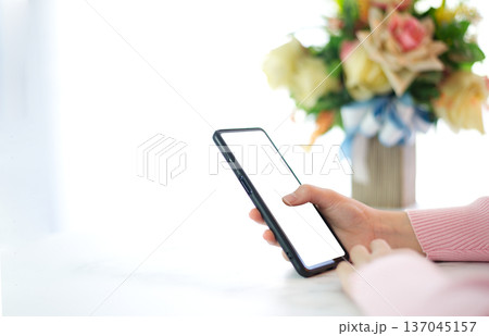 Hands of woman in pink shirt holding smart phone with white screen on white table top decorated with bouquet of flowers in a bright room background 137045157