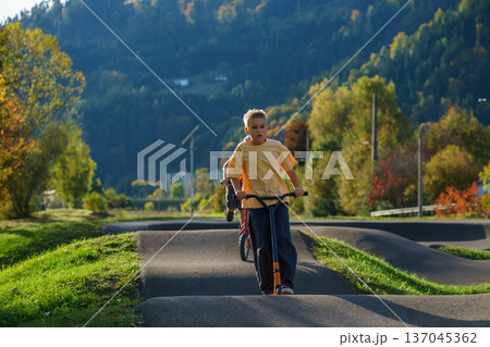 Child riding scooter on asphalt pump track with scenic hills and trees behind. Sporty lifestyle, outdoor adventure, freedom and active youth concept. Child riding scooter on asphalt pump track with scenic hills and trees behind. Sporty lifestyle, outdoor adventure, freedom and active youth concept. 137045362