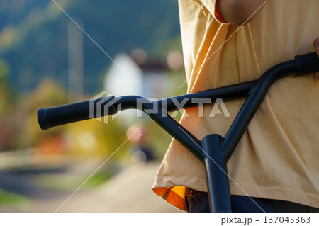 Close view of scooter handlebar held by child in warm evening sunlight. Outdoor activity, mobility, modern transport and carefree childhood moment. Close view of scooter handlebar held by child in warm evening sunlight. Outdoor activity, mobility, modern transport and carefree childhood moment. 137045363