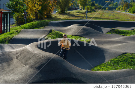 Focused boy riding scooter through curved asphalt pump track in urban park. Active childhood, balance training,outdoor recreation and sporty lifestyle 137045365