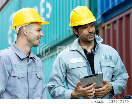Two construction workers wearing yellow safety helmets discussing project details with digital tablet at shipping container yard, industrial teamwork and logistics management concept 137047179