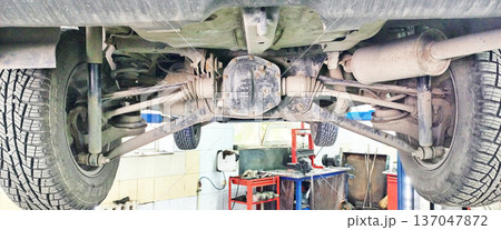 Underside view of a vehicle raised on a lift in a repair shop, showing the rear differential, suspension components, and winter tires Underside view of a vehicle raised on a lift in a repair shop, showing the rear differential, suspension components, and winter tires 137047872