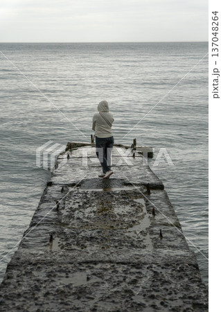 Person standing on concrete pier facing Black Sea, overcast sky, muted tones and calm water create minimalist coastal scene with focus on solitude and travel experience. Tourism, wellness 137048264