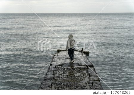 Person standing on concrete pier facing Black Sea, overcast sky, muted tones and calm water create minimalist coastal scene with focus on solitude and travel experience. Tourism, wellness 137048265