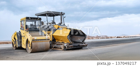 Heavy equipment-a roller and paver-works smoothly, laying asphalt, while construction crews lay the foundation for a new road against a picturesque cloudy backdrop 137049217