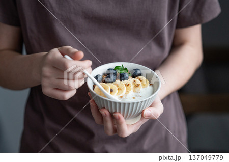 Woman holding bowl of granola with greek yogurt, apple, blueberries and spoon in hands Woman holding bowl of granola with greek yogurt, apple, blueberries and spoon in hands 137049779