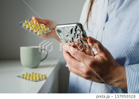 Woman taking fish oil omega 3 capsules for woman health and reading instruction for medication in the internet on smart phone close up. Supplements and vitamins, self treatment. Woman taking fish oil omega 3 capsules for woman health and reading instruction for medication in the internet on smart phone close up. Supplements and vitamins, self treatment. 137050328