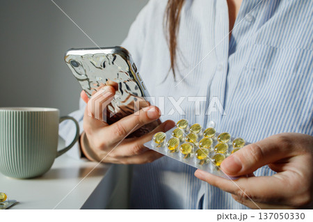 Woman taking fish oil omega 3 capsules for woman health and reading instruction for medication in the internet on smart phone close up. Supplements and vitamins, self treatment. Woman taking fish oil omega 3 capsules for woman health and reading instruction for medication in the internet on smart phone close up. Supplements and vitamins, self treatment. 137050330