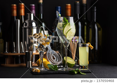 Bar composition featuring a variety of cocktails: martini with olives, citrus mint highball, and limoncello shot against wine bottles, shaker, strainer and fresh ingredients Bar composition featuring a variety of cocktails: martini with olives, citrus mint highball, and limoncello shot against wine bottles, shaker, strainer and fresh ingredients 137050468