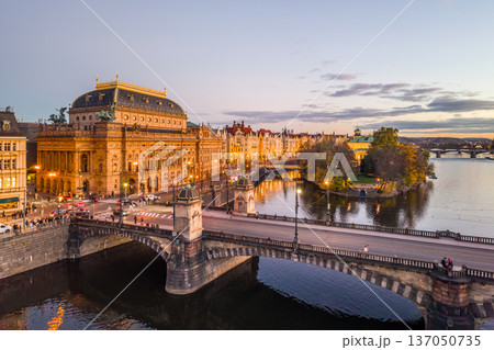 Aerial view of the National Theatre in Prague during the evening. The theatre is lit up, and the surrounding embankment and bridges are visible. 137050735