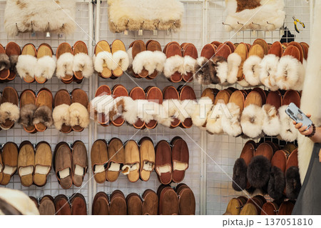 Sheepskin slippers displayed on market stall rack in retail market stall, various colors and sizes. Craft sheepskin slippers sale, winter footwear shopping, handmade leather goods Sheepskin slippers displayed on market stall rack in retail market stall, various colors and sizes. Craft sheepskin slippers sale, winter footwear shopping, handmade leather goods 137051810