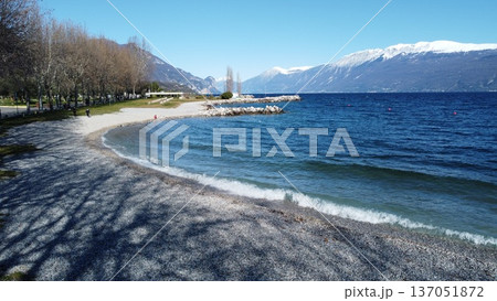 Pebble beach in Toscolano Maderno on Lake Garda with snow-capped mountains. 137051872