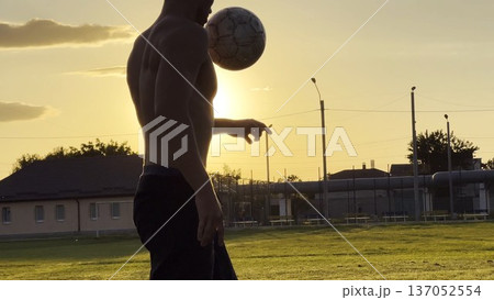 Young man juggling soccer ball on stadium at sunset. Professional footballer kicking ball at green field. Sportsman practicing tricks at meadow with sunlight at background. Freestyle football Young man juggling soccer ball on stadium at sunset. Professional footballer kicking ball at green field. Sportsman practicing tricks at meadow with sunlight at background. Freestyle football 137052554