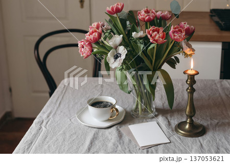 Spring flowers in glass vase on wooden table. Blurred kitchen background. Red tulips, eucalyptus, white anemone flowers bouquet. Candleholder, candle, greeting card mockup. Scandinavian interior. Spring flowers in glass vase on wooden table. Blurred kitchen background. Red tulips, eucalyptus, white anemone flowers bouquet. Candleholder, candle, greeting card mockup. Scandinavian interior. 137053621