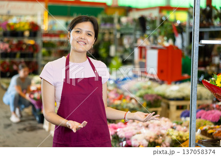 Portrait of smiling young woman gardener standing at greenhouse with flowers on background Portrait of smiling young woman gardener standing at greenhouse with flowers on background 137058374