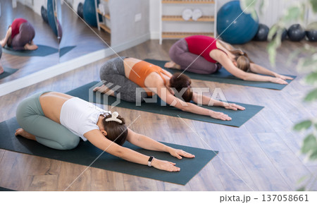 Sporty female lying on yoga mat and doing exercise yoga together with group of people in fitness studio Sporty female lying on yoga mat and doing exercise yoga together with group of people in fitness studio 137058661