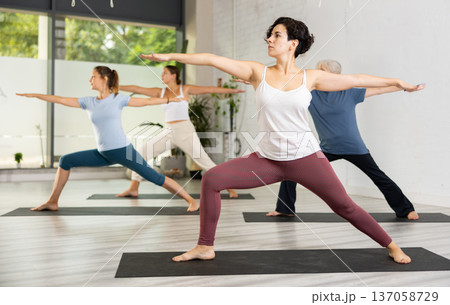 Active young Latin woman practicing Virabhadrasana 2 Warrior pose, indoor class with women, studio background Active young Latin woman practicing Virabhadrasana 2 Warrior pose, indoor class with women, studio background 137058729