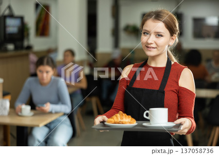 Female cafe employee carries coffee and croissants on tray Female cafe employee carries coffee and croissants on tray 137058736