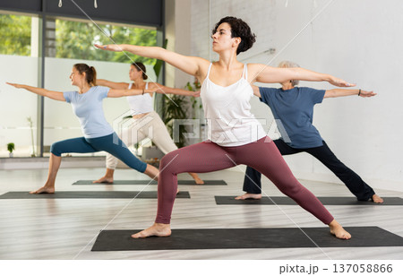 Active young Latin woman practicing Virabhadrasana 2 Warrior pose, indoor class with women, studio background 137058866